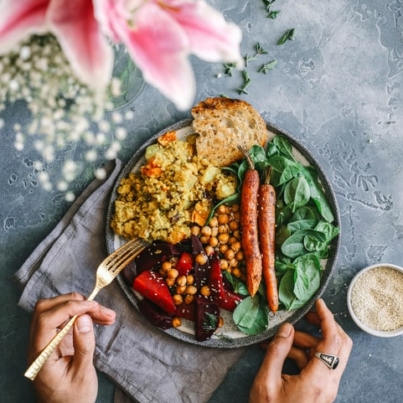 hands holding a fork near a bowl of food with greens, carrots, legumes, and bread