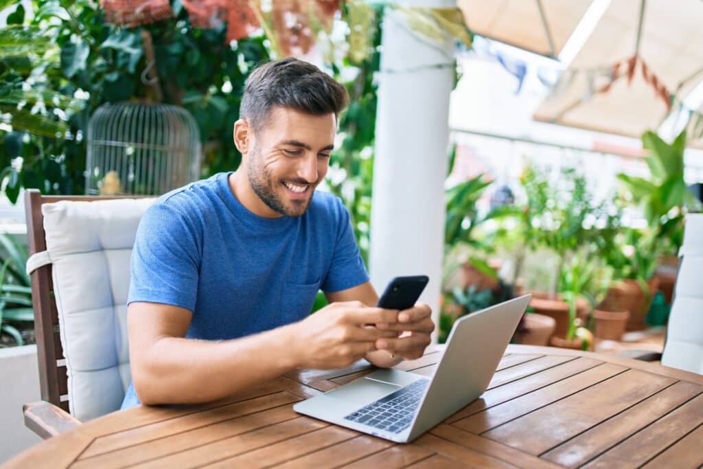 man sitting at an outdoor table holding his phone with a laptop in front of him