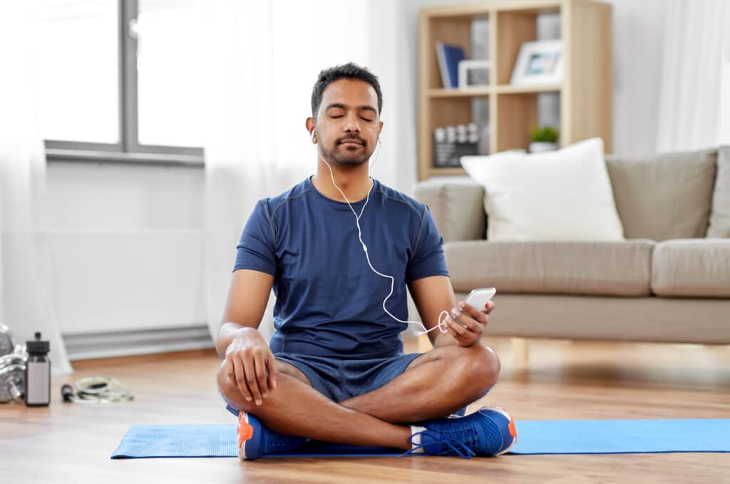 Man in athletic wear sitting on yoga mat with phone in hand and headphones in meditating